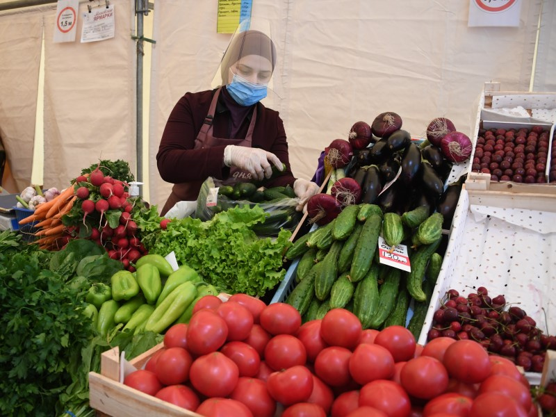 На фото продавщица на рынке, на переднем плане овощи: перец, лук, огурцы, салат, редис. Сама продавщица в маске, защитном экране и перчатках